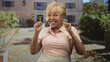 © Krakenimages.com - Senior hispanic woman wearing yellow eyeglasses claps hands and smiles, hands near chest, standing in a sunlit stone building courtyard with fountain; joy gratitude.