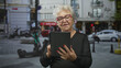 © Krakenimages.com - Senior woman with white hair and glasses tapping a tablet screen with finger while standing on a busy urban street near scooters and parked cars and storefronts; curiosity.