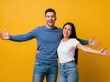 © Vasiliy - Happy young couple smiling with open arms in a welcoming gesture. Cheerful man and woman in casual clothes embracing against a yellow background with copy space