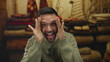 © Krakenimages.com - Hispanic man making funny faces on a carpet shop indoors, showcasing playful expressions with a joyful demeanor, surrounded by colorful textiles and warm lighting.