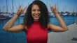 © Krakenimages.com - Woman with curly hair smiling in red shirt makes rock horns gesture at a port pier; joyful celebration.