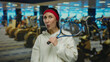 © Krakenimages.com - Woman in gym holding racket, smiling energetically in a sports center setting with gym equipment visible, wearing red headband and white sweatshirt.