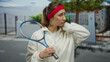 © Krakenimages.com - Woman standing outdoors holding racket looking thoughtful wearing casual urban sportswear with tree background capturing city street ambiance and youthful vitality.