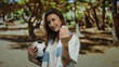 © Krakenimages.com - Woman outdoors in a park holding a soccer ball with blue scarf making inviting gesture with a hand and smiling warmly, surrounded by blurred trees in the background.