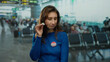 © Krakenimages.com - Young woman wearing vote badge sitting in airport terminal, gesturing with her hand against a busy indoor backdrop, expressing a message of civic duty and participation.