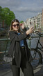 © Krakenimages.com - Woman taking selfie by amsterdam canal with phone, showcasing urban lifestyle with bicycle in background and vibrant cityscape on a sunny day.