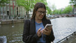 © Krakenimages.com - Woman smiling while using smartphone by amsterdam canal in spring day.