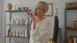 © Krakenimages.com - Woman holds smartphone to her lips in a pottery studio surrounded by clay vases and shelves; concentration.
