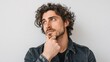 © mila103 - A man with curly hair looks upwards with a thoughtful expression. He holds his chin with one hand while standing against a simple wall inside a room.
