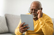 © Prostock-studio - An elderly man is focused on reading content on a tablet while seated on a couch. He adjusts his glasses to improve his view. The living room is bright and comfortable.