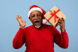 © Prostock-studio - An older man wearing a red sweater and a Santa hat smiles broadly while holding a gift. He appears to be enjoying a festive moment during the holiday season.