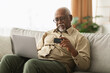 © Prostock-studio - A senior man sits comfortably on a couch, focused on his laptop while holding a credit card. Sunlight fills the room, creating a warm and inviting atmosphere.