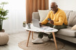 © Prostock-studio - An older man sits on a light-colored couch, smiling as he holds a credit card and looks at his laptop. Natural light fills the room, creating a cozy atmosphere.