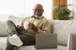 © Prostock-studio - A senior man sits on a sofa in a cozy living room, smiling as he drinks coffee and looks at his laptop. Natural light fills the space, creating a warm atmosphere.