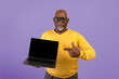 © Prostock-studio - A senior man stands against a plain purple background holding a laptop. He is smiling and pointing at the screen while wearing a yellow sweater and eyeglasses.