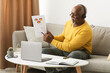 © Prostock-studio - A senior man wearing headphones explains a colorful chart while seated on a couch in his living room. A laptop and notebook are on a table in front of him.