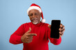 © Prostock-studio - An elderly man wearing a red sweater and a Santa hat is smiling while holding up a smartphone. He points at the phone with his other hand. The background is light blue and festive.