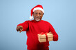 © Prostock-studio - An elderly man stands against a light blue background. He wears a red sweater and Santa hat. He holds a gift box with a ribbon and points outward, smiling joyfully.