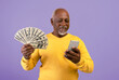 © Prostock-studio - A man stands with a big smile, holding a stack of cash in one hand and a smartphone in the other. He is focused on the screen of the phone. The background is purple.