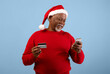 © Prostock-studio - A man in a red sweater and Santa hat looks at his phone while holding a credit card. He appears to be shopping online during the holiday season.
