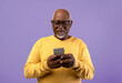 © Prostock-studio - A man with gray hair and glasses holds a smartphone in his hands, focusing on the screen. He wears a yellow sweater and stands in front of a purple backdrop.