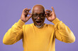 © Prostock-studio - A senior man with gray hair adjusts his large glasses with both hands. He shows a surprised expression. The background is purple. He wears a yellow long-sleeve shirt.