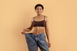 © Anastasiya - Smiling young black woman holding oversized jeans to show weightloss results. Toned abs, healthy glow and positive energy ideal for fitness, transformation and beauty care campaigns