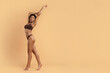 © Anastasiya - Young black woman stretching upward in brown underwear on beige background. Highlights flexibility, body toning, beauty-care routines and weightloss focused wellness lifestyle, copy space
