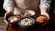 © photo for everything - A person presents a culinary arrangement of small bowls filled with diverse dishes on a wooden tray. Dishes include fish, tofu, rice, kimchi, and soup, showcasing the art of presentation.