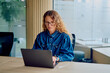 © we.bond.creations - Woman wearing glasses and a denim shirt concentrating on her laptop at a desk in a contemporary office environment