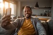 © ttonaorh - Smiling man sitting on couch taking a selfie with his smartphone in cozy ambient lighting. Evening home relaxation and connection.