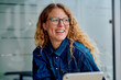 © we.bond.creations - Woman smiling brightly with glasses and curly hair, enjoying a moment while working in a modern office with her tablet
