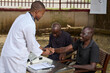 © The Yudel Media - A doctor shakes hands with a smiling adult man seated at a table while another man sits beside them during an outdoor medical checkup.