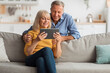 © Prostock-studio - A happy couple sits on a comfortable couch, smiling as they watch a video on a tablet. The cozy living room features soft furnishings and warm decor.