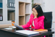 © R Photography - An Asian businesswoman sits at her office desk, appearing stressed and fatigued. She experiences headaches, migraines, neck shoulder pain, and numb hands from long working hours and office syndrome