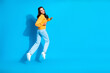 © deagreez - Young mixed race woman jumping in a yellow sweater and light blue jeans against a vibrant blue studio backdrop