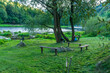 © Volha - Quiet picnic area with wooden benches near a riverbank surrounded by trees and grass. Outdoor recreation space, camping mood, nature relaxation, and peaceful countryside leisure concept.