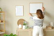 © Pixel-Shot - Young woman hanging blank frame on beige wall at home, back view