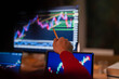 © Studio Marmellata - A person in a red shirt uses a pencil to point at a stock market graph on a computer screen, with other screens showing similar data in the foreground.