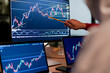 © Studio Marmellata - A person points at a stock market chart on a computer screen, analyzing financial data with multiple monitors displaying trading graphs.