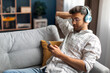 © Home-stock - Relaxed Indian man chilling on couch at home, using wireless headphones and cellphone, watching video content online, copy space