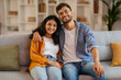 © Home-stock - Portrait of positive young Indian spouses embracing, sitting on couch at home and smiling at camera