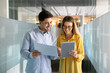 © fizkes - Happy young coworkers comparing sales reports, analyzing paper document and electronic content on tablet, standing in office hall, corridor, talking, discussing financial analytic data