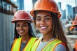© Aleksandra - Two young women wearing orange safety helmets and yellow reflective vests smiling at a construction site with urban buildings in the background
