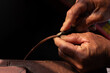 © Nguyen Tuan - Close up of artisan's hand using a cutting tool to trim a brown leather strip. Handmade craftsmanship and precise leather working process.