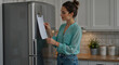 © Vyatcheslav - Young woman looking at a paper on a modern kitchen refrigerator. Organizing home tasks and daily routine. Blank sheet for copy space