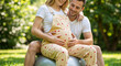 © Vyatcheslav - An affectionate expectant couple touching a pregnant belly outdoors. A smiling man and woman sitting together on an exercise ball in a park. Maternity and family concept