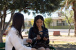 © Rangel - Young women sitting on a park bench, sharing headphones and a moment of connection, representing friendship, relaxed communication, and modern leisure activities on a sunny day