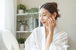 © KMPZZZ - Facial beauty skin care, smile of pretty asian young woman in bathrobe looking at mirror, hand applying moisturizer lotion on her face, putting cream treatment before makeup cosmetic routine at home.