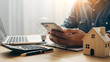 © Mounya - A person using a smartphone on a desk with a laptop, calculator, house model, and lightbulb, symbolizing financial planning or real estate investment